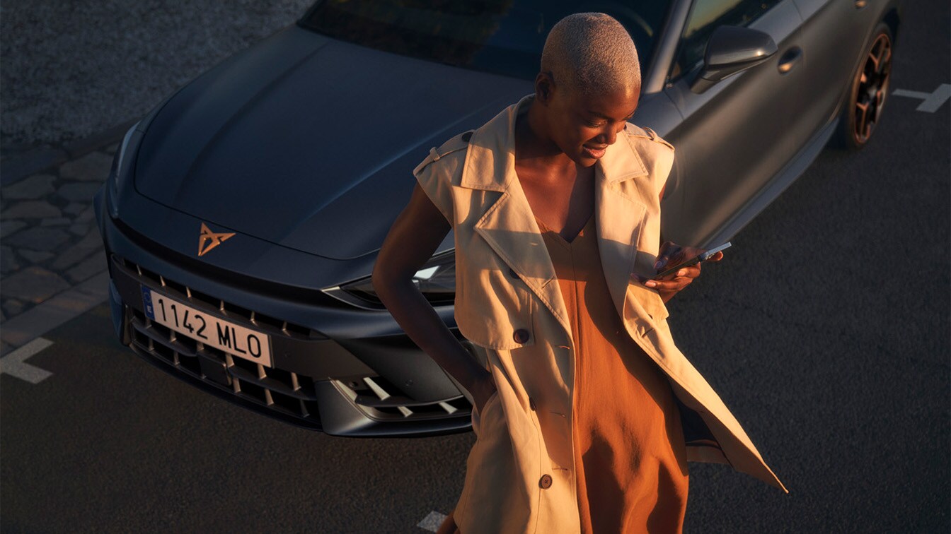 A woman with short-cropped hair wearing a beige sleeveless trench coat over a rust-colored dress, looking at a smartphone and standing in front of a CUPRA car, captured in warm golden hour light.