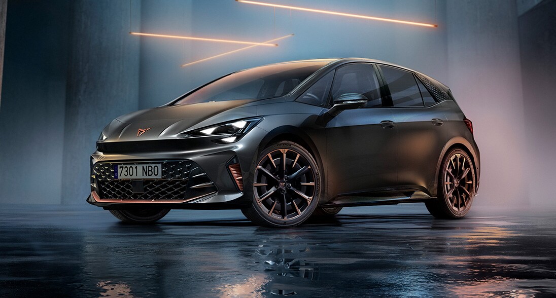 A CUPRA Born parked in a modern concrete architectural setting with a wet, highly reflective floor. The vehicle features illuminated sharp headlights and multi-spoke alloy wheels, highlighted by a series of glowing orange geometric light bars suspended overhead.