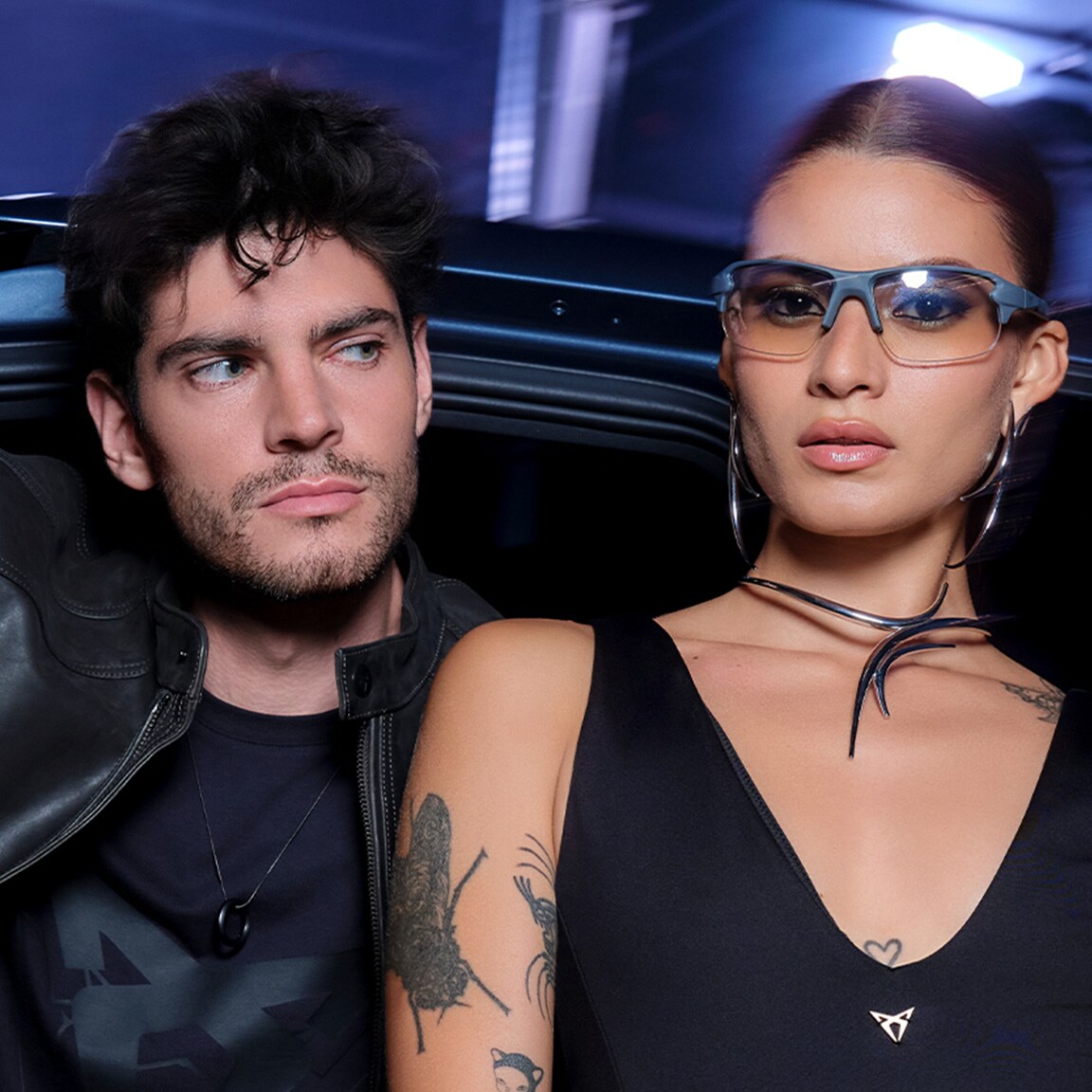A man in a black leather jacket leans out of a dark CUPRA car door next to a woman wearing sunglasses and large hoop earrings. The woman has a tattoo on her arm and a metallic choker. The scene is dark, lit by interior and ambient blue light.