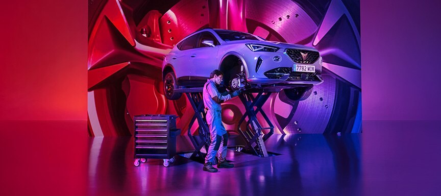 A mechanic works on the front wheel area of a CUPRA car elevated on a scissor lift. The stylized studio setting features a highly reflective floor, a rolling tool cabinet, and a large background screen displaying an oversized, vibrant graphic of a car wheel, all bathed in pink, red, and blue gradient lighting.
