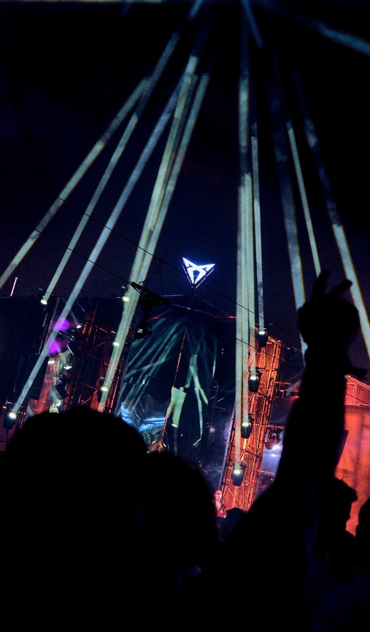 Perspective from a crowd at a night music festival under fanned searchlights from a large stage with a central glowing CUPRA inverted triangle logo.