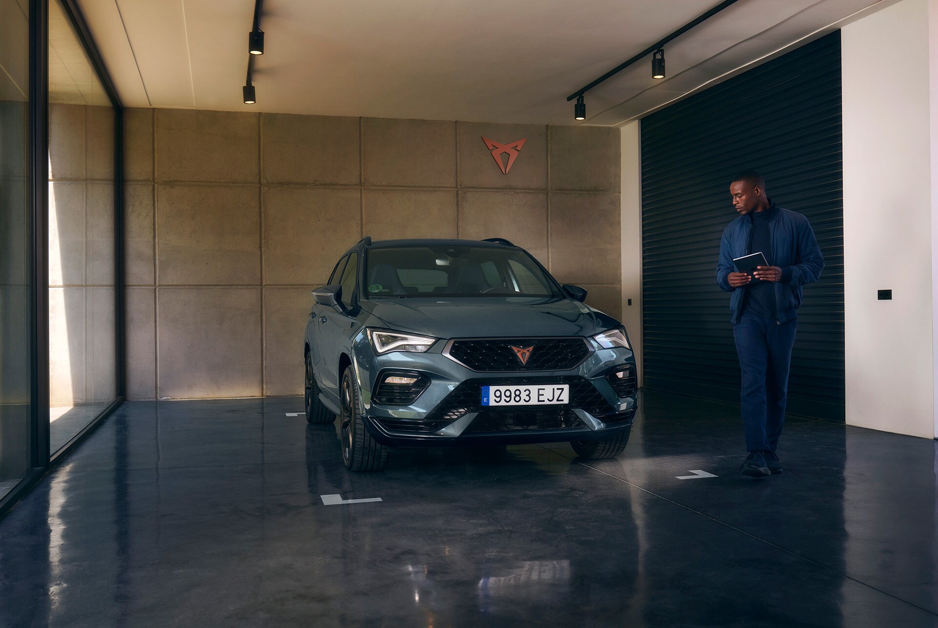 A dark gray CUPRA car is parked inside a modern garage with concrete walls and a polished dark floor. A man is walking toward the car, holding a tablet. A CUPRA logo is mounted on the wall.