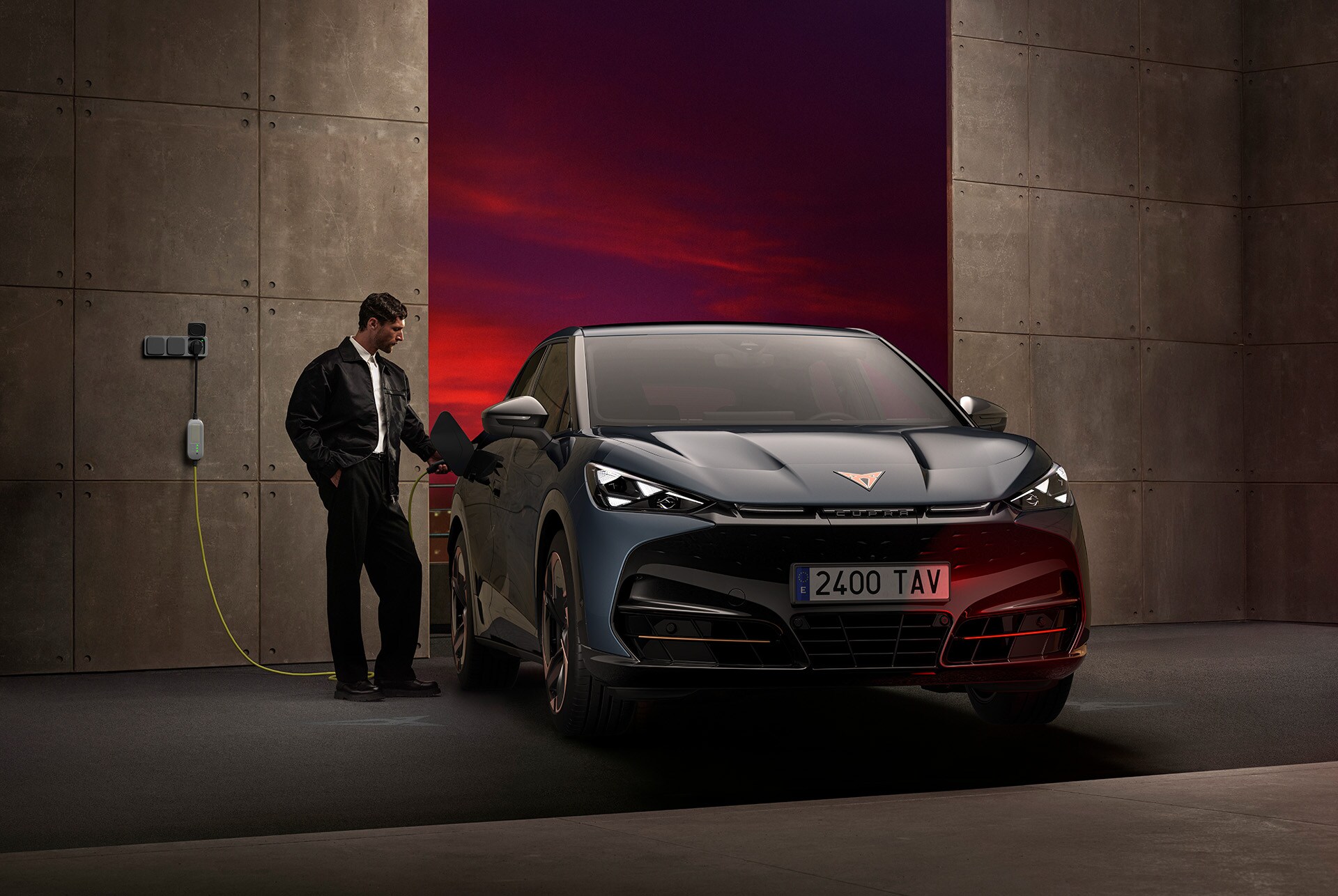 A man is standing next to the front of a dark CUPRA car that is plugged into a wall charger. He is interacting with the car's charging port, and the car is parked in a dark garage with a vibrant red and purple background.