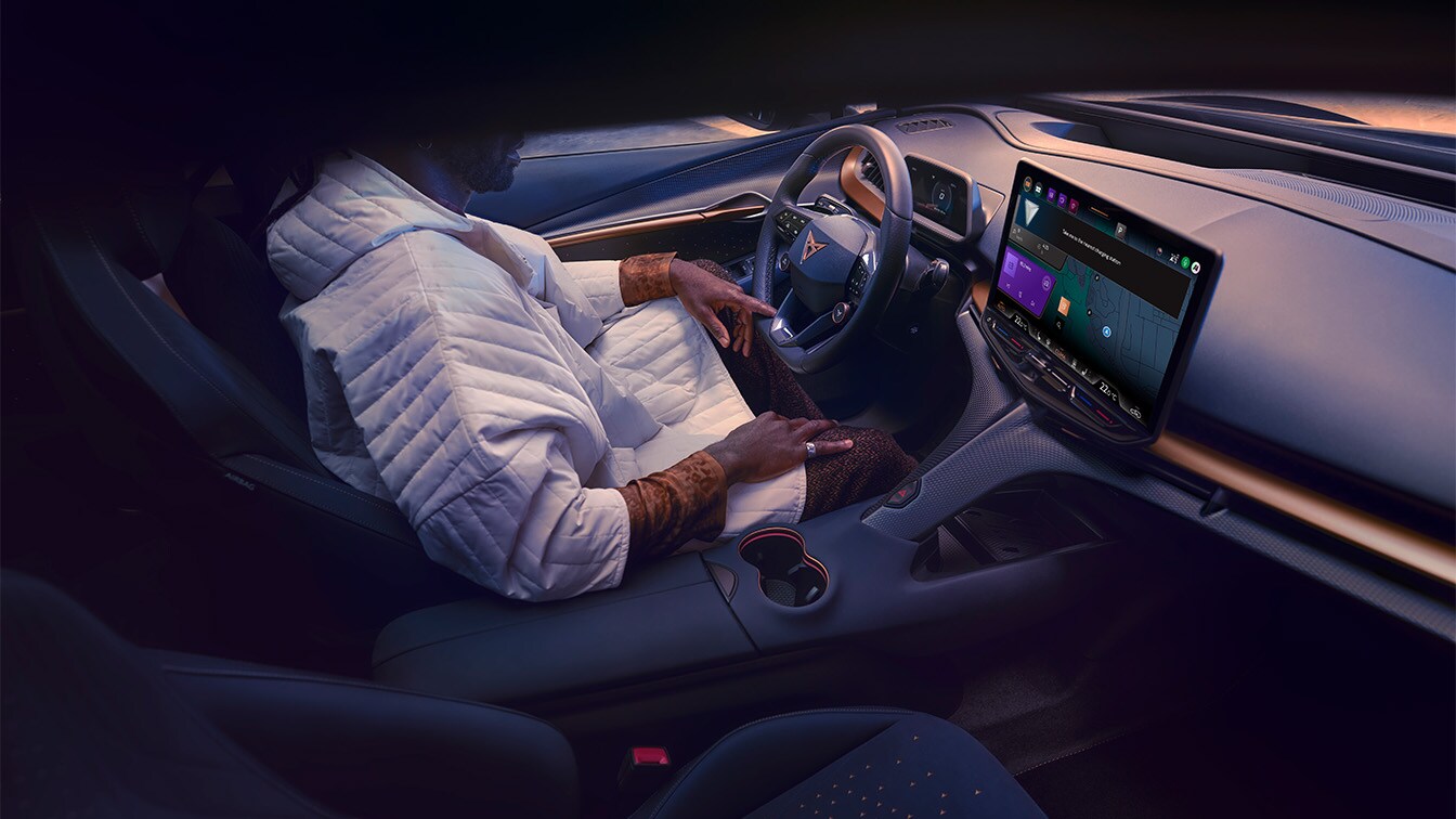 Interior view of the CUPRA Tavascan from the passenger side, showing a driver sitting in the front seat with the central infotainment touchscreen displaying the digital assistant interface. 