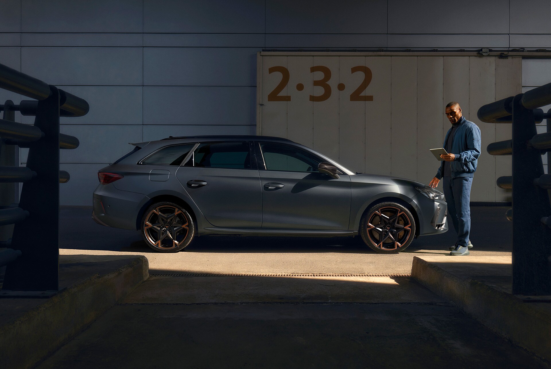 A dark gray CUPRA wagon is parked in a partially shaded, industrial area. A man in a blue jacket stands at the front of the car, looking down at a tablet.