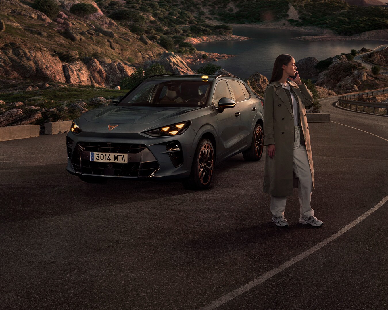 A gray CUPRA car is parked on the side of a road at dusk. A person is sitting in the driver's seat with the door open, while two other people stand outside, one on the left and one on the right. The sky has strong orange and blue hues.